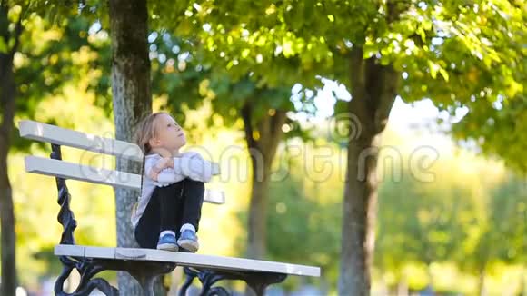 可爱的小女孩在美丽的秋日户外秋天可爱的孩子坐在户外公园的长凳上视频的预览图