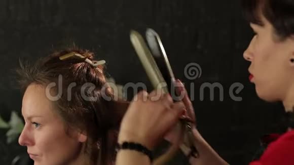 女理发师用卷发熨斗在红色头发上做卷发美容院视频的预览图
