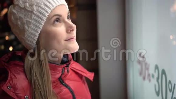 美丽迷人的年轻女人看着商店的橱窗红色的冬衣销售标志晚上好视频的预览图