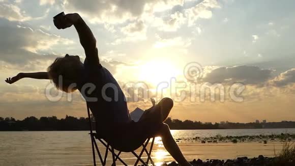 快乐的人坐在河堤上邋遢的夕阳下伸展视频的预览图