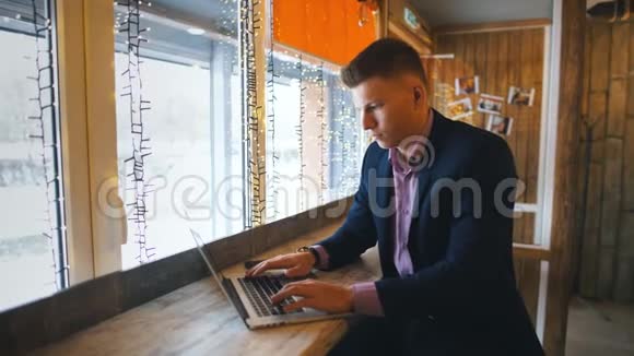 在咖啡馆工作穿着智能休闲装看着笔记本电脑坐在创意办公室靠窗的开朗的年轻人视频的预览图