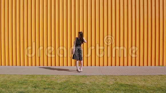 在一个阳光明媚的日子里穿着黑白裙子的美丽年轻女子沿着黄色的墙走了一半视频的预览图