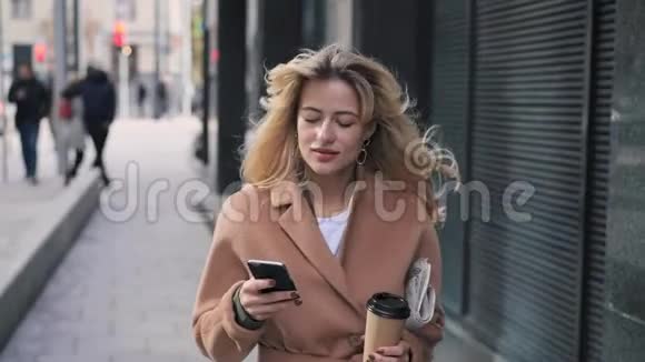 迷人的金发女人带着咖啡散步用她的智能手机视频的预览图