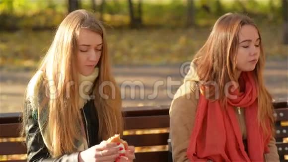 两个美丽的女孩在公园吃快餐在阳光明媚的秋天坐在长凳上吃午饭的学生视频的预览图