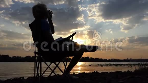 年轻人在日落时用记事本在折叠椅上喝咖啡视频的预览图