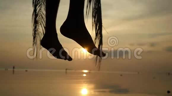 年轻人坐在棕榈树上在美丽的夕阳下在热带海滩挥舞着雄腿的剪影慢动作视频的预览图
