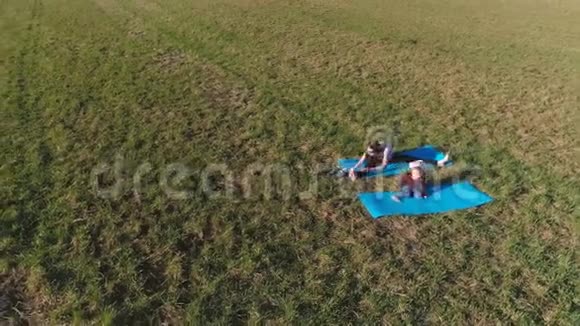 在温暖的夏日里妈妈和她的小女儿在田野上的垫子上伸展空中全景视频的预览图
