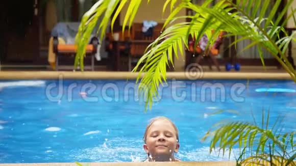 兴奋的快乐男孩在雨中用一棵棕榈树的树枝在一个有蓝色水的游泳池里游泳慢动作1920x1080视频的预览图