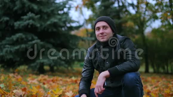 男人在镜头里打招呼挥动双手微笑着坐在落叶背景下的草地上视频的预览图