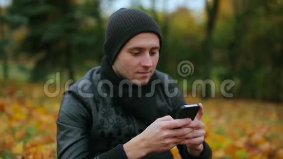 秋天一个男人坐在公园里玩智能手机打字和聊天短信的肖像特写视频的预览图