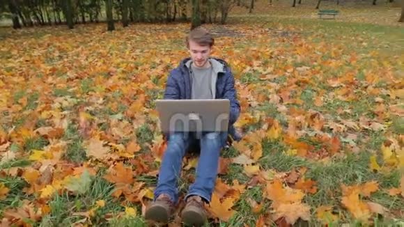 年轻的男性it创业程序员使用笔记本电脑在秋天公园的落叶地毯上工作视频的预览图