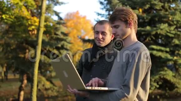 两个微笑的创业商人在秋天的公园里谈论分享笔记本电脑视频的预览图
