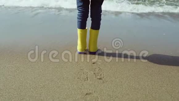 小女孩穿着亮黄色胶靴的特写镜头孩子站在沙滩上脚印在沙滩上视频的预览图