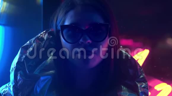年轻的时尚少女戴着毛皮眼镜吹着泡泡糖上面有街上霓虹灯蓝色的粉红色标志美丽极了视频的预览图