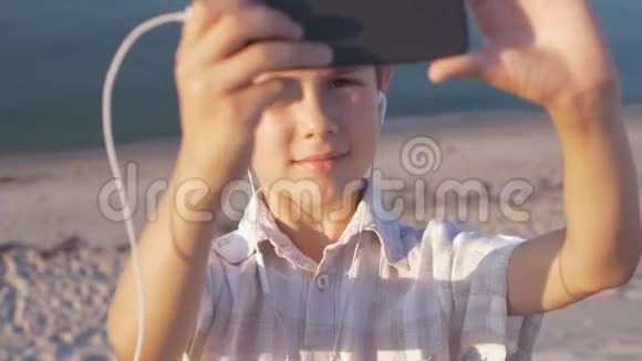 可爱的男孩在海滩上自拍用智能手机在沙滩上拍摄海洋背景视频的预览图