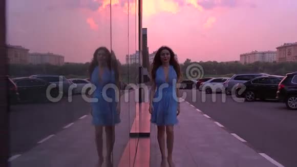 苗条的黑发女孩穿着蓝色连衣裙在粉红色日落天空的街道上散步4K视频视频的预览图