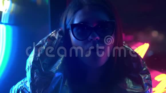 年轻的时尚少女戴着毛皮眼镜吹着泡泡糖上面有街上霓虹灯蓝色的粉红色标志美丽极了视频的预览图