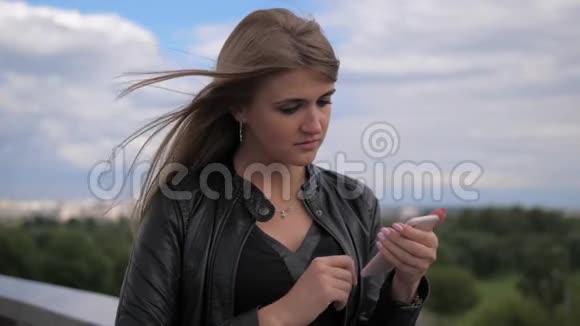 漂亮的金发美女正在看手机触摸屏幕在街上视频的预览图