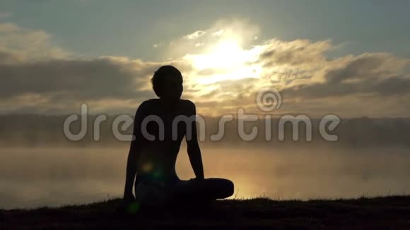 年轻人坐在湖畔在日落时慢动作练习瑜伽视频的预览图