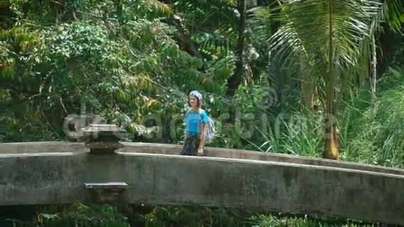 女孩头上戴着绷带背包穿过热带丛林中的旧石桥游客步行视频的预览图