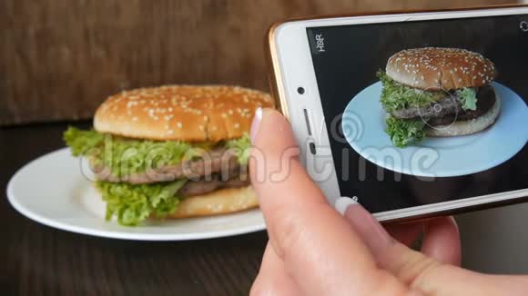 女性手在智能手机上制作一个漂亮的多汁汉堡在时尚的木制上放生菜叶视频的预览图