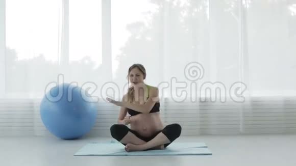 美丽的怀孕少女大肚腩正在健身房里跳舞坐在地板上莲花的位置视频的预览图
