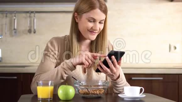 美丽的年轻迷人的女人吃早餐和使用她的智能手机喝果汁视频的预览图