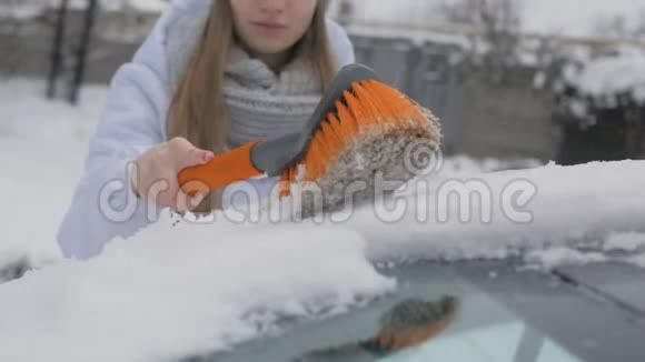 年轻漂亮的女孩用刷子擦车上的雪视频的预览图