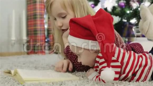 戴着圣诞帽的女孩和男孩在圣诞树前看书视频的预览图