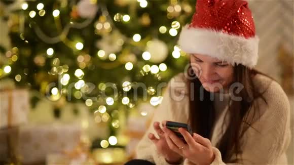 圣诞老人帽子里的布鲁内特在手机上打字在背景圣诞树上微笑圣诞帽女孩用小玩意视频的预览图
