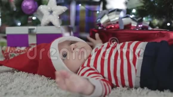 戴着圣诞帽的婴儿躺在圣诞树旁带着礼物视频的预览图