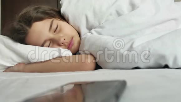 小女孩在床上睡觉小女孩醒来看着智能手机闹钟在室内的生活方式视频的预览图