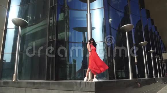 漂亮的女孩穿着红色连衣裙戴着蓝色眼镜在建筑背景上戴着太阳镜的布鲁内特对着相机微笑黄色视频的预览图