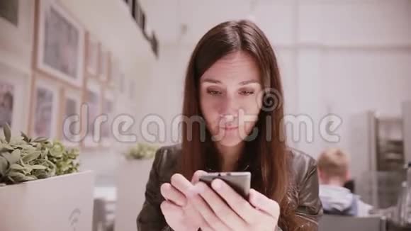 漂亮的年轻女人黑发和眼睛穿着皮夹克使用她的智能手机灯光室墙上的图片视频的预览图