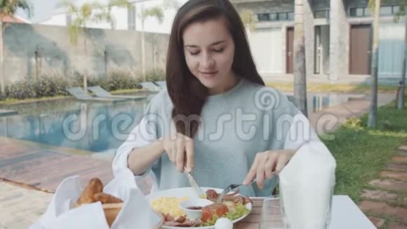 女人吃早餐时吃炒鸡蛋香肠靠近豪华别墅的游泳池视频的预览图
