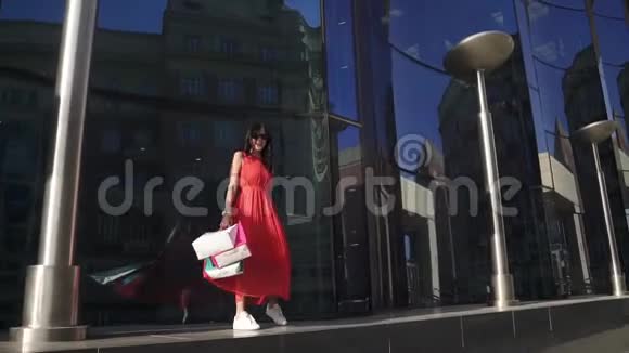 一个穿着红色连衣裙的顽皮女孩走在现代蓝色玻璃建筑旁边反射和阳光视频的预览图