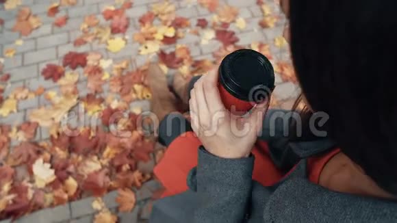 秋天美丽的枫叶坐在城市公园喝咖啡的年轻时尚女性的俯视图视频的预览图