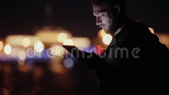 帅哥微笑点头晚上用他的平板电脑然后走开城市灯光模糊侧视现代技术视频的预览图