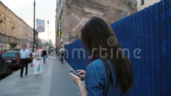 年轻女子站在街道上的一个蓝色栅栏附近女士用智能手机把头转向相机慢节奏视频的预览图