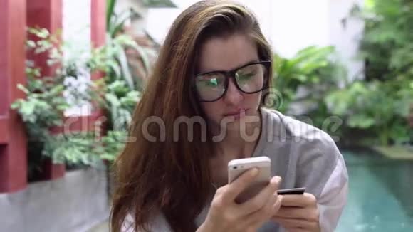 戴眼镜的女人在智能手机上使用网上银行信用卡在热带国家的游泳池附近视频的预览图