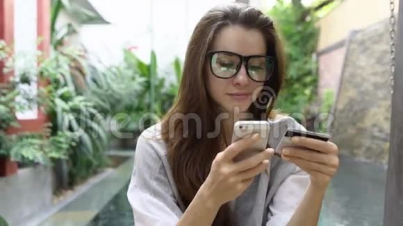 戴眼镜的女人在智能手机上使用网上银行信用卡在热带国家的游泳池附近视频的预览图