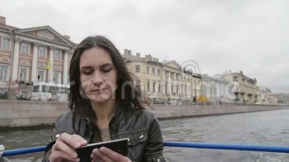 年轻女子在河边旅游拍照使用智能手机风吹头发圣彼得堡的城市景色慢节奏视频的预览图