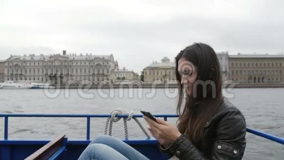 美丽的黑发女人用她的智能手机坐在河边的公共汽车上休息她高兴地笑了俄罗斯圣彼得堡视频的预览图
