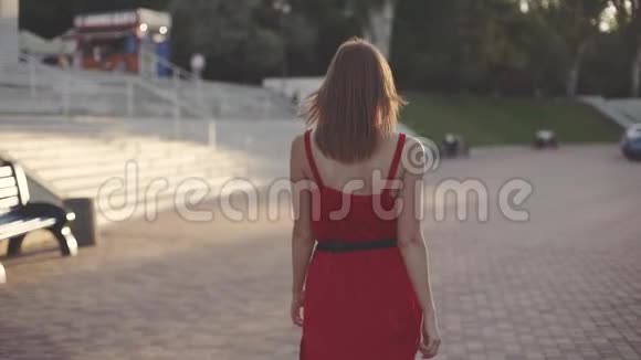 美丽的年轻红发女子穿着红色的裙子走在街上在日落时玩得很开心视频的预览图