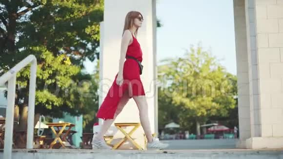 美丽的年轻红发女子穿着红色的裙子走在街上在日落时玩得很开心视频的预览图