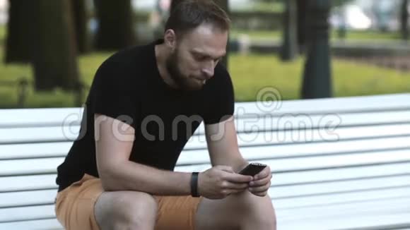 在公园里坐在长凳上用智能手机发送短信一个笑容满面的年轻人视频的预览图