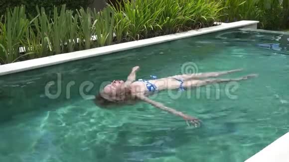 在热带度假胜地的棕榈树下比基尼有一头长发的年轻苗条女子在游泳池里游泳视频的预览图