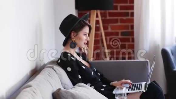 穿着黑色帽子和裙子的女孩坐在沙发上拿着笔记本电脑视频的预览图