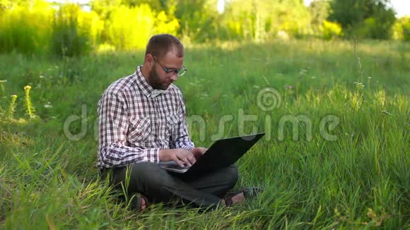 戴眼镜的年轻人坐在草坪上在膝上打字视频的预览图