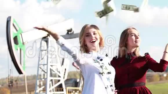 两个年轻快乐的女人在油田油泵的背景上扔美元视频的预览图